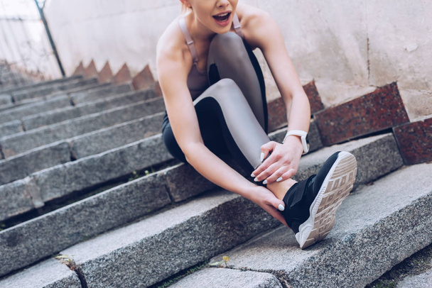young sportswoman sitting on stairs and suffring from hurt while touching injured leg - Foto, Imagem