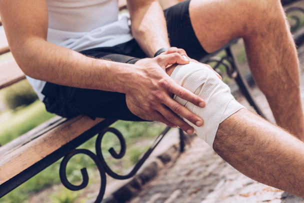 cropped shot of sportsman sitting on bench and touching elastic bandage on injured knee - Фото, изображение