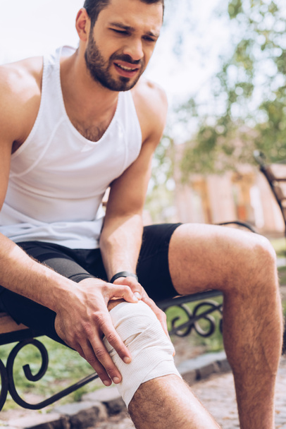 upset sportsman sitting on bench and touching elastic bandage on injured knee - Fotó, kép