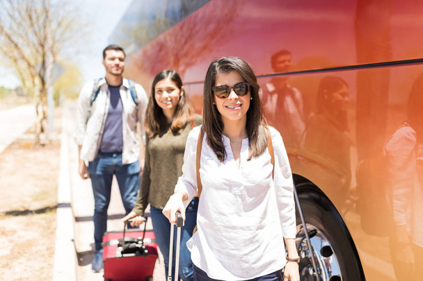 Retrato de una hermosa mujer joven con gafas de sol mientras está de pie junto a los turistas y autobús de viaje
 - Foto, imagen