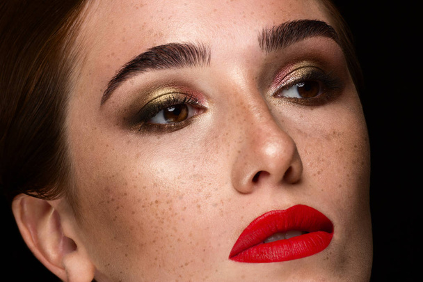Close-up of a red-haired girl with freckles and fashionable evening make-up. Sensual plump lips with red lipstick, dark shadows on the eyelids and thick eyebrows. The fatal image of a beautiful woman. - Fotografie, Obrázek