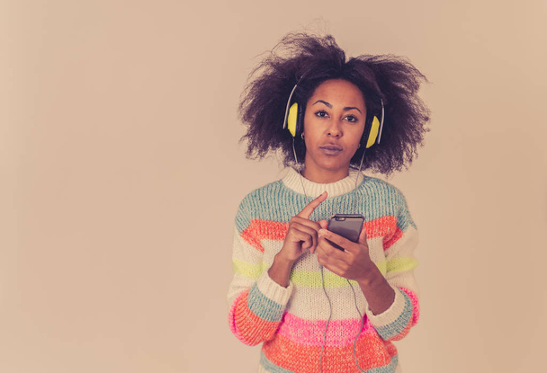 Πολύχρωμο πορτρέτο της πανέμορφης και ευτυχισμένη αφρικανική Αμερικανίδα γυναίκα ακούγοντας έξυπνη τηλεφωνική μουσική εφαρμογή στα ακουστικά τραγουδώντας και χορεύοντας σε ουδέτερο φόντο. Στους ανθρώπους μουσική τεχνολογία και την ευτυχία. - Φωτογραφία, εικόνα