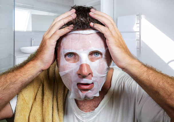 young scared and surprised man at home using beauty paper facial mask cleansing doing anti aging facial treatment looking himself weird and funny in toilet mirror - Foto, afbeelding