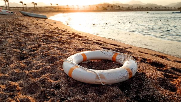 Closeup image of white plastic life saving ring lying on the sandy sea beach against beutiful sunset over the water - Photo, Image