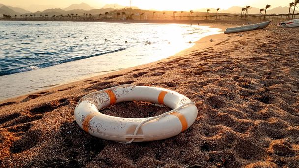 Κοντινό πλάνο με λευκό πλαστικό δαχτυλίδι εξοικονόμησης ζωής που βρίσκεται στην αμμώδη παραλία με όμορφο ηλιοβασίλεμα πάνω από το νερό - Φωτογραφία, εικόνα