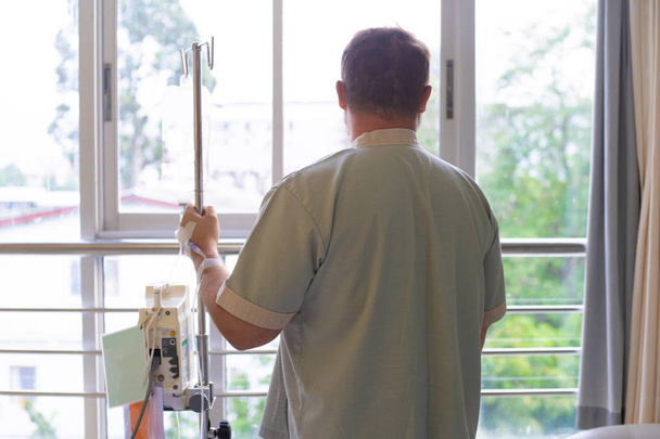 ασθενής με ένα σταγονόμετρο κοιτάζει έξω από το παράθυρο του νοσοκομείου και χαμόγελα. πίσω όψη. Έννοια της υγειονομικής περίθαλψης. - Φωτογραφία, εικόνα