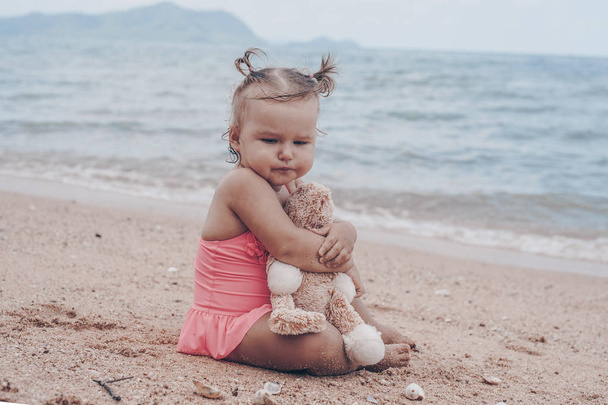 töprengő kislány teddy medve átölelve és keres el a tengerparton ülve. - Fotó, kép