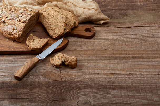 čerstvý řezený chléb na sekací desce v blízkosti hadříku a nože na dřevěném stole - Fotografie, Obrázek
