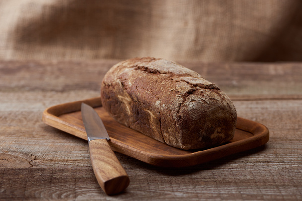 świeży brązowy chleb bochenek na deski do krojenia w pobliżu noża na drewnianym stole - Zdjęcie, obraz