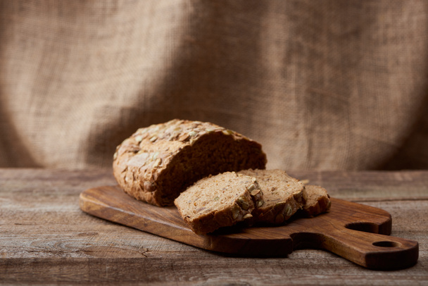 delicious sliced brown bread on wooden chopping board with sackcloth on background - Фото, изображение