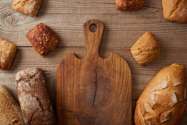 вид сверху на свежие домашние буханки хлеба и булочек возле пустой доски на деревянном деревенском столе
 - Фото, изображение