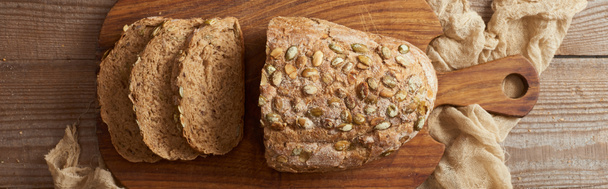pohled na čerstvý domácí řezený chléb s osivem na dřevěné sekací desce v blízkosti hadříku, panoramatický záběr - Fotografie, Obrázek