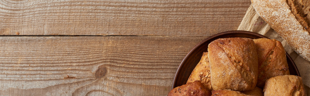 Draufsicht auf frische Brotlaibe und Brötchen in Schüssel auf Tuch auf Holztisch, Panoramaaufnahme - Foto, Bild