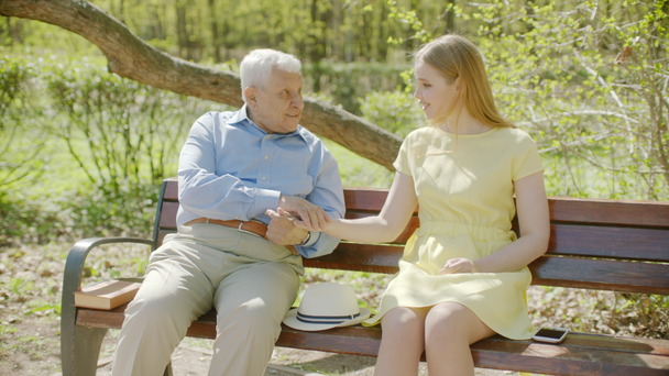 Young woman and her grandfather having a talk on the bench in the park on summer day - Video, Çekim