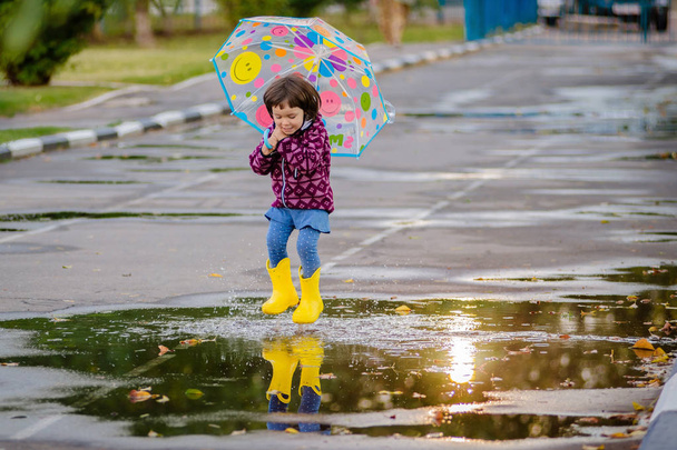 Niño divertido feliz con paraguas multicolor saltando charcos en botas de goma y riendo
 - Foto, imagen