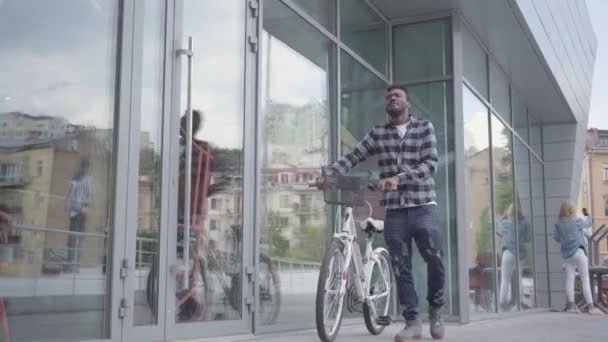 Portrait confident handsome African American man standing with bicycle against the background of urban architecture. The guy is resting in good weather outdoors - Filmmaterial, Video