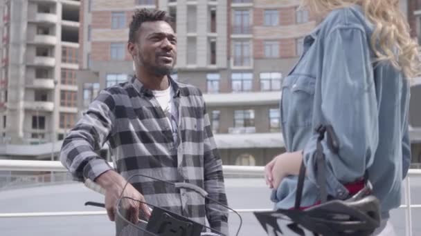 Portrait handsome African American man standing with bicycle against the background of urban architecture talking with an cute attractive blond woman. Couple of friends chatting outdoors - Video