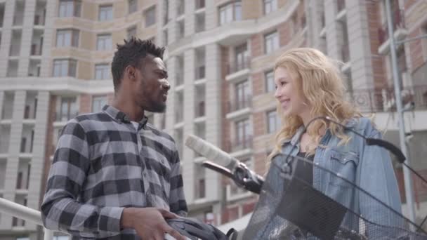 Portrait of handsome African American man with bicycle against the background of urban architecture talking with attractive woman. Couple of friends chatting outdoors. International friendship - Materiał filmowy, wideo