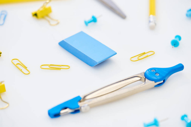 selective focus of blue compasses, eraser and paper clips mixed with yellow paper clips and pencil on white background - Photo, Image