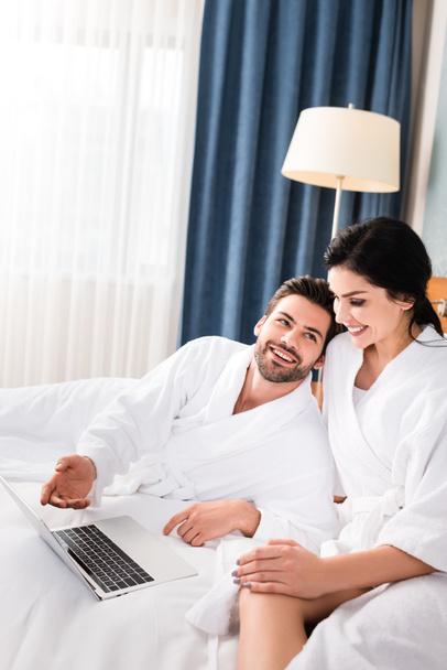 cheerful man gesturing near laptop and looking at happy brunette woman in hotel room  - Fotó, kép