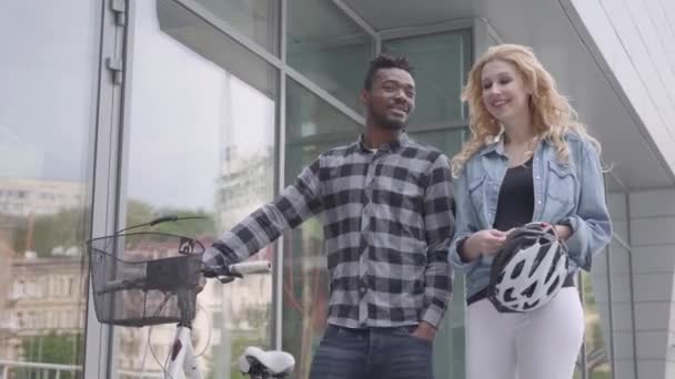 Handsome confident African American man in casual clothes leading his bicycle near the shop windows in city talking with Caucasian girlfriend. The international couple spending time together outdoors - Video