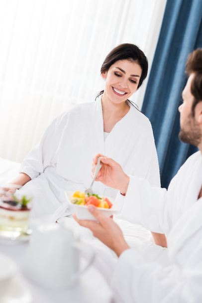 selective focus of happy woman looking at bearded man holding fork near tasty fruit salad  - 写真・画像