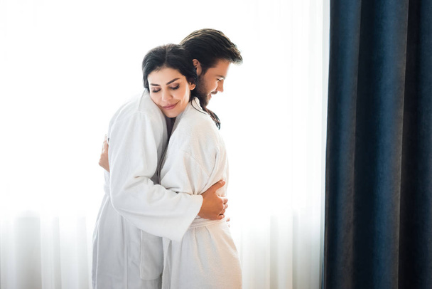 cheerful bearded man hugging attractive woman in hotel room  - Photo, Image