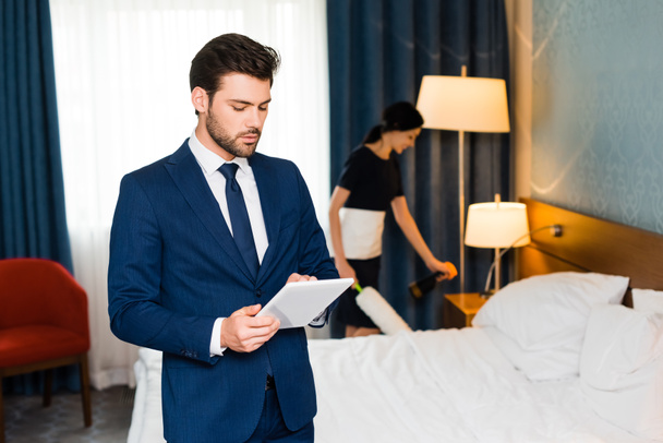selektywne skupienie przystojnego człowieka za pomocą cyfrowego tabletu w pobliżu pokojówki w pokoju hotelowym  - Zdjęcie, obraz