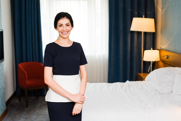 χαρούμενη υπηρέτρια χαμογελά ενώ κοιτάζει την κάμερα στο δωμάτιο του ξενοδοχείου  - Φωτογραφία, εικόνα