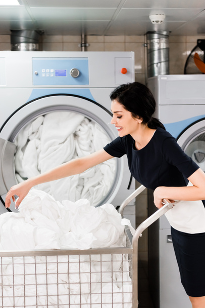 cheerful maid touching bed sheets while standing near washing machines in laundry  - Φωτογραφία, εικόνα