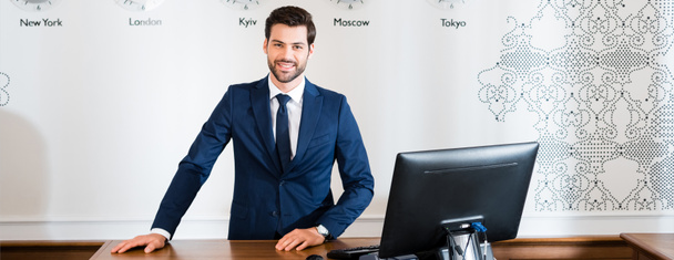 panoramic shot of happy receptionist standing at reception desk in hotel  - Photo, Image