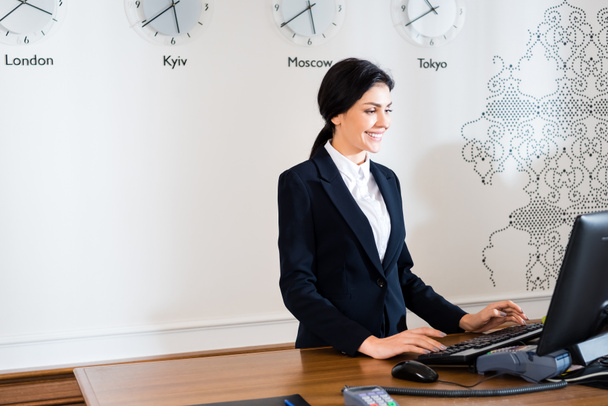 happy brunette receptionist in formal wear standing near computer monitor in hotel  - Photo, Image