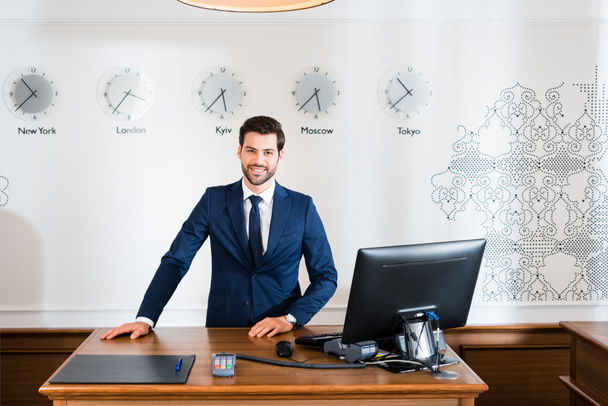 cheerful receptionist in suit standing near computer monitor in hotel  - Photo, Image