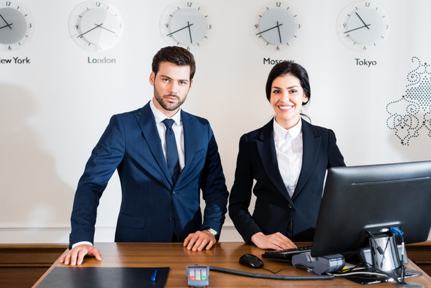 cheerful woman near serious receptionist in suit standing at reception desk  - Photo, Image