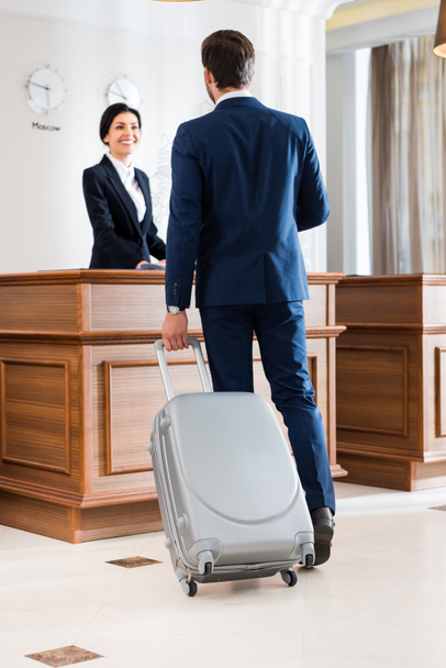 επιλεκτική εστίαση του επιχειρηματία με βαλίτσα στέκεται κοντά όμορφη ρεσεψιονίστ  - Φωτογραφία, εικόνα