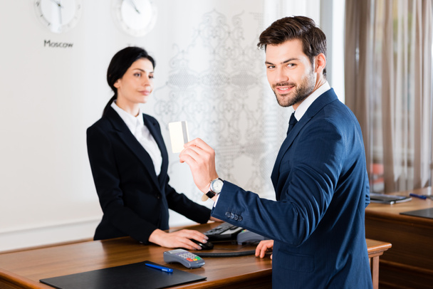 selective focus of cheerful businessman holding credit card near serious receptionist  - Photo, Image