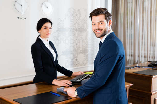 selective focus of happy businessman holding credit card near serious receptionist  - Photo, Image