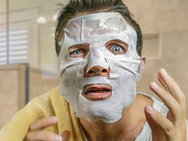 young funny and messy man applying beauty facial mask looking to the mirror surprised and horrified in skin care aging concept and male cosmetic face treatment - Fotó, kép