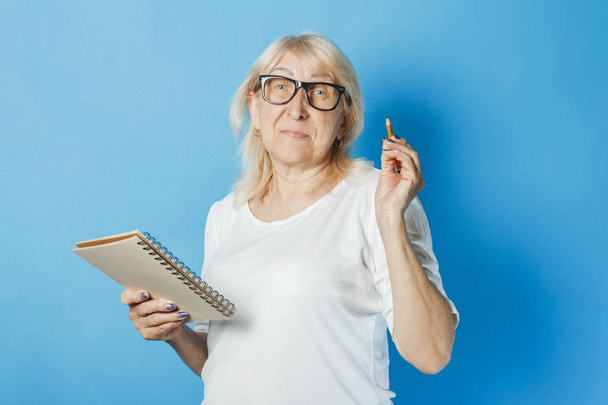 Eine alte Dame mit Brille hält vor blauem Hintergrund ein Notizbuch und einen Stift in der Hand. Konzeptnotizen, Kreuzworträtsel, Sudoku, Stapelliste, schlechtes Gedächtnis - Foto, Bild