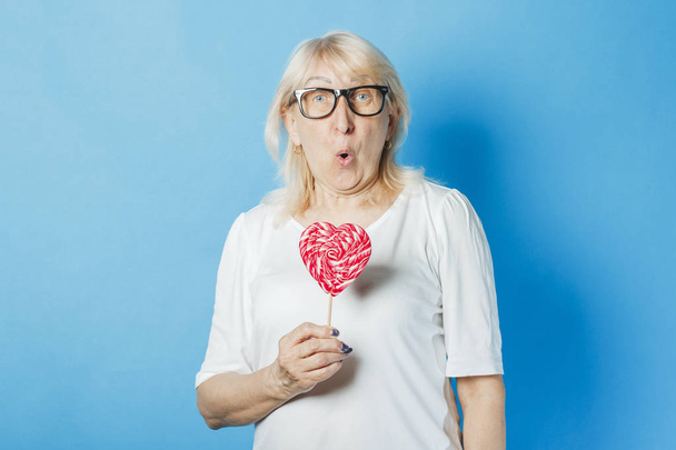 Öreg hölgy szemüveg gazdaság szív alakú nyalóka a kék háttér. Fogalma édességek, szerelem, Valentin-nap - Fotó, kép