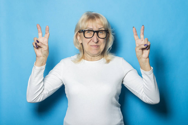 eine alte Frau mit Brille und glücklichem Gesicht macht eine Geste mit den Händen vor blauem Hintergrund. Konzept Glück, gute Laune, alles ist gut - Foto, Bild