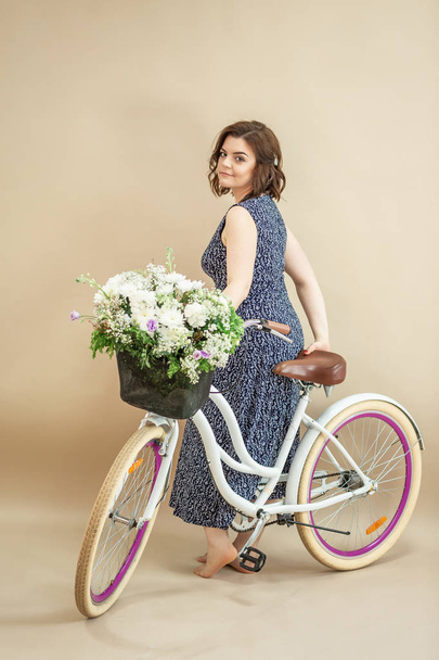 Hermosa chica sonriente está montando la bicicleta con la cesta llena de flores aisladas sobre fondo beige - Foto, imagen