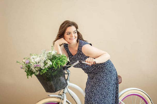 Mujer joven romántica está de pie con su bicicleta y una cesta con flores silvestres. Verano. aislado sobre fondo beige - Foto, imagen