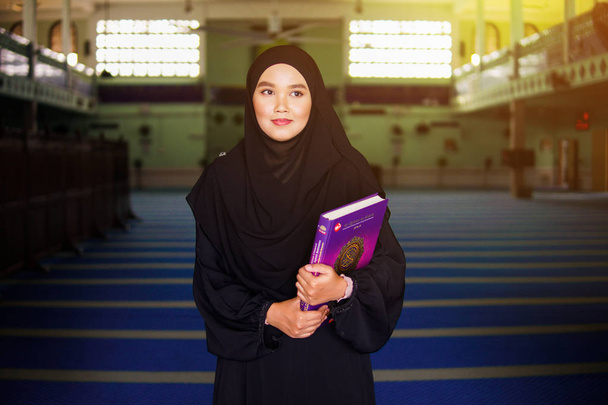 Молодая мусульманка в черном платье из абайи держит в себе Священный Коран в мечети
 - Фото, изображение