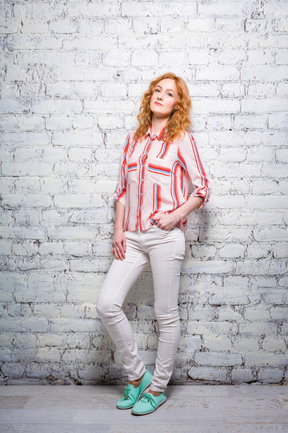 Full-length portrait A young beautiful Caucasian woman student stands near a white brick wall. Girl with red long curly hair with freckles on her face in bright summer clothes - Foto, Imagem