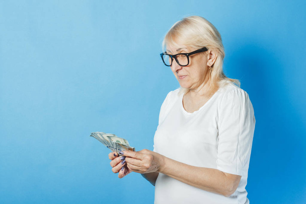 Stará dáma s brýlemi drží peníze v dlaních na modrém pozadí. Koncept bohatství, výher, úspor, loterie, půjčky - Fotografie, Obrázek