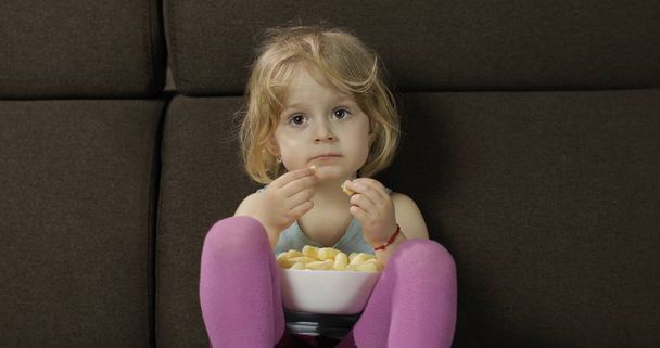 Girl sitting on sofa and eating corn puffs. Child taste puffcorns - Foto, afbeelding