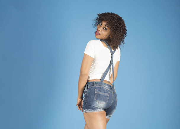 Lady with makeup looks back. Woman with african appearance in denim clothes looks gorgeous and stylish on blue background. Lady in modern clothes as short denim overalls. Fashion concept - Photo, Image