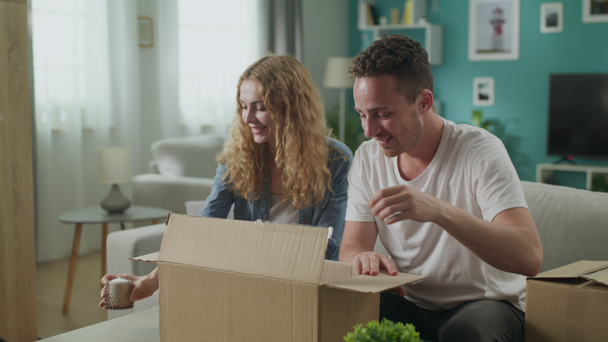 Happy couple sitting on couch in living room open unpacking cardboard box parcel - Séquence, vidéo