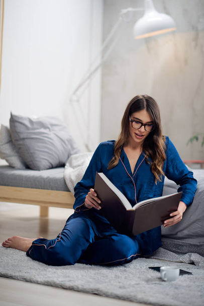 Young gorgeous Caucasian brunette in pajamas sitting on the floor in bedroom and reading book in the morning. - Foto, imagen